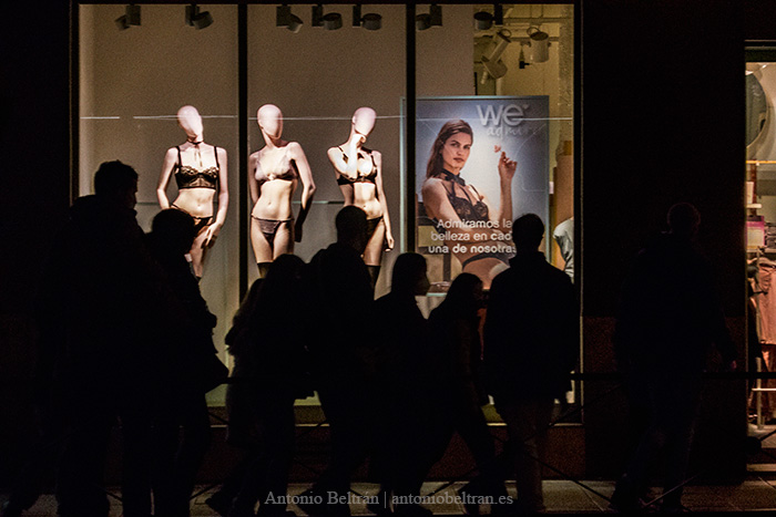 un grupo de personas miran un escaparate de maniquíes femeninos en ropa interior. Es de noche.